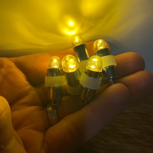 Può includere: Un gruppo di piccole lampadine a LED gialle. Ogni lampadina ha una parte superiore trasparente e arrotondata e una base argentata, tenute insieme da un elastico bianco. Le lampadine emettono una luce gialla brillante e calda.