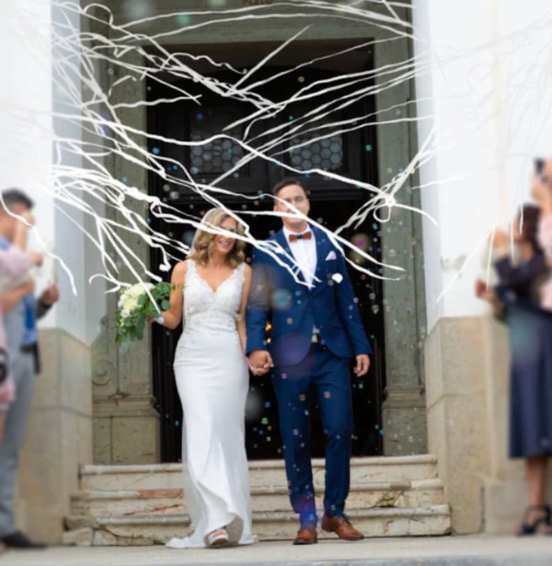 May include: A newly married couple exits a building, holding hands. The bride wears a white wedding dress with lace detailing, and the groom wears a blue suit with a bow tie. White streamers and bubbles float around them, celebrating their wedding.
