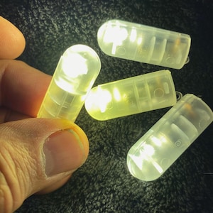 May include: Four small, clear, capsule-shaped LED lights are illuminated. One is held by a person's fingers. The lights are a soft yellow color and appear to be battery-operated. The plastic casings have a ridged design.