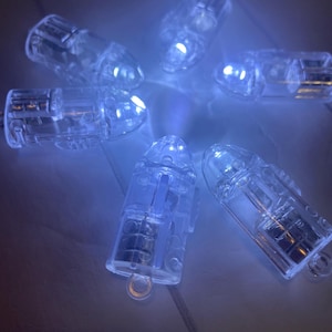 Może przedstawiać: Zbiór małych, przezroczystych, kulistych diod LED emitujących delikatną niebieską poświatę. Każda lampka ma małą pętlę u podstawy. Światła są rozmieszczone na powierzchni, tworząc subtelne, ambientowe oświetlenie.