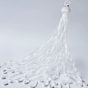 May include: A white decorative object with a circular top and long, flowing ribbons. The ribbons cascade downwards, with small, white, circular elements attached at intervals. The object is set against a plain white background.