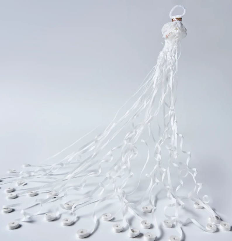 May include: A white decorative object with a circular top and long, flowing ribbons. The ribbons cascade downwards, with small, white, circular elements attached at intervals. The object is set against a plain white background.