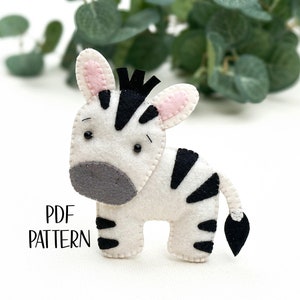 Puede incluir: Un cebra de fieltro blanco y negro con orejas rosas y ojos negros. El cebra está de pie sobre una superficie blanca. El texto "PDF PATTERN" es visible en la esquina inferior izquierda de la imagen.