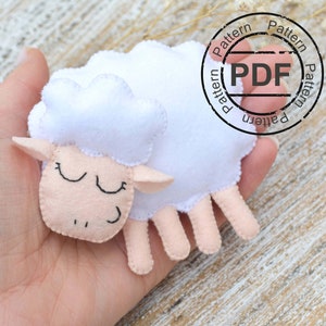 May include: A white felt sheep with pink felt details and black embroidered eyes. The sheep is lying down with its eyes closed, as if sleeping. The sheep is being held in a person's hand.