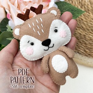 Può includere: Un cervo in feltro fatto a mano con dettagli marroni, bianchi e rosa. Il cervo ha piccole corna marroni, un naso nero e dettagli cuciti. Il testo "PDF PATTERN + SVG template" è visibile. Questo è probabilmente un modello artigianale.