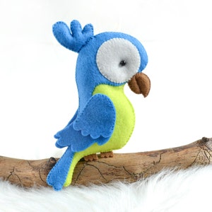 May include: A blue and green felt parrot toy perched on a branch. The parrot has a white eye and a brown beak.