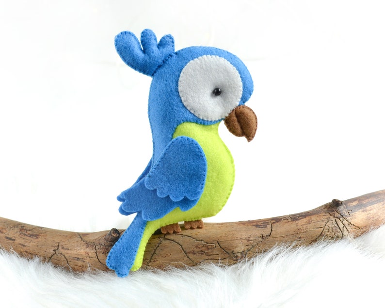 May include: A blue and green felt parrot toy perched on a branch. The parrot has a white eye and a brown beak.