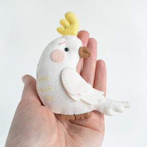 May include: A white felt cockatoo bird with a yellow crest, brown eyes, and pink cheeks. The bird is sitting in a hand.