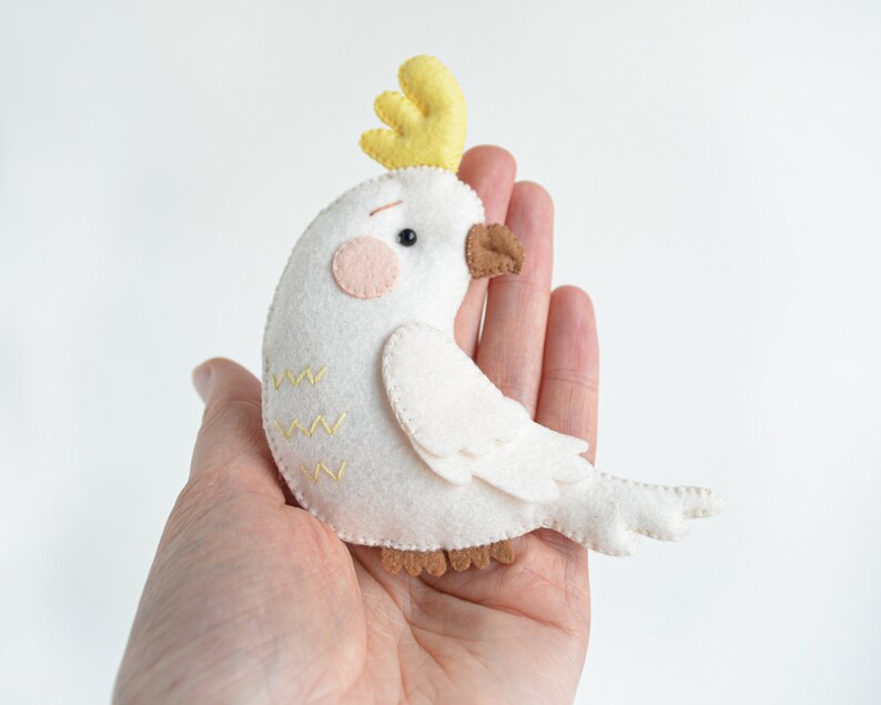 May include: A white felt cockatoo bird with a yellow crest, brown eyes, and pink cheeks. The bird is sitting in a hand.
