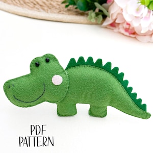 May include: A green felt alligator toy with black eyes and a pink felt nose. The alligator has a jagged green felt back fin. The toy is on a white surface. PDF PATTERN.