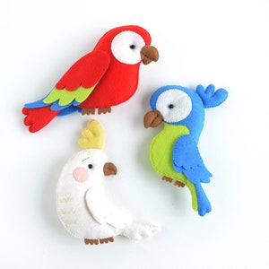 May include: Three felt bird toys. A red parrot with green and blue feathers, a blue parrot with green feathers, and a white cockatoo with a yellow crest.