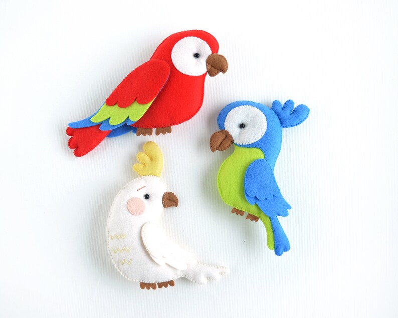 May include: Three felt bird toys. A red parrot with green and blue feathers, a blue parrot with green feathers, and a white cockatoo with a yellow crest.