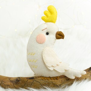 May include: A white felt cockatoo with a yellow crest, brown beak, and pink cheeks. The cockatoo is perched on a branch.