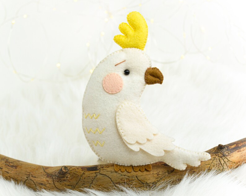 May include: A white felt cockatoo with a yellow crest, brown beak, and pink cheeks. The cockatoo is perched on a branch.