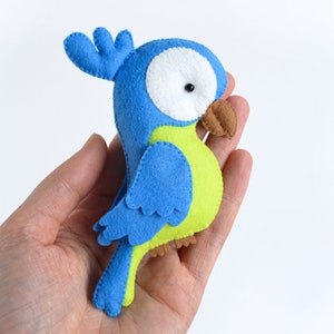 May include: A blue and green felt parrot toy with a white eye and a brown beak. The toy is held in a person's hand.