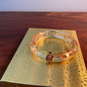 May include: A clear resin bangle bracelet with embedded seashells. The shells are various shades of brown, orange, and white.