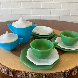 Puede incluir: Un juego de té vintage con una tetera y una cremaera turquesa con tapas blancas, y tazas y platillos verdes. El juego se exhibe en una bandeja de madera, creando una estética encantadora y nostálgica.