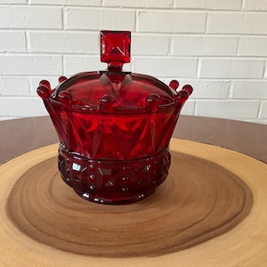 Puede incluir: Un tarro de vidrio rojo rubí en forma de corona con tapa y asa cuadrada. El tarro tiene una base decorativa y está sobre una superficie de madera. El fondo es una pared de ladrillos blancos.