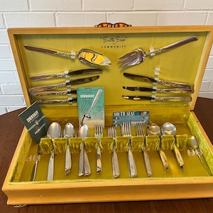 May include: A wooden box with a yellow velvet interior containing a set of Community "South Seas" silverware. The silverware includes knives, forks, spoons, and serving pieces. The box is open and the silverware is arranged in the velvet lining.