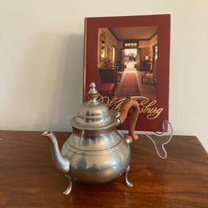 May include: A silver teapot with a wooden handle and a wooden knob on the lid. The teapot is sitting on a wooden table with a book titled "Williamsburg" in the background.