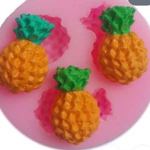 Könnte beinhalten: Rosa Silikonform mit drei Ananasformen. Die Ananas sind gelb mit grünen Spitzen.