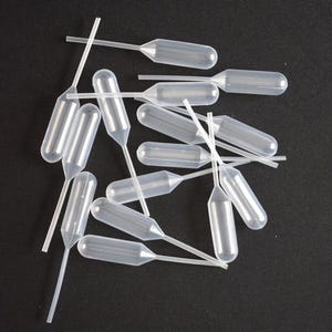 Puede incluir: Un conjunto de pipetas de plástico transparente, con extremos en forma de bulbo y tallos finos y alargados. Las pipetas están dispuestas sobre un fondo oscuro, destacando su diseño transparente. Herramientas de laboratorio.