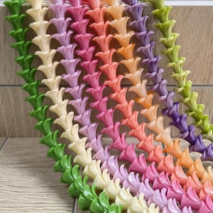 May include: A collection of seven vertical strands of small, trumpet-shaped flower beads arranged in a gradient of colors including green, cream, lavender, pink, peach, orange, and purple, each featuring a smooth, glossy finish and uniform shape.