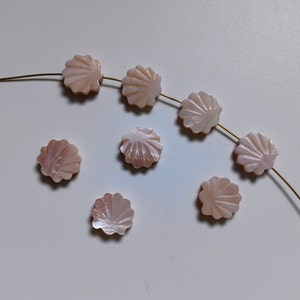 May include: A strand of pink shell beads with a scalloped edge. The beads are strung on a gold wire and are arranged in a curved line. There are also several loose beads on the white surface.