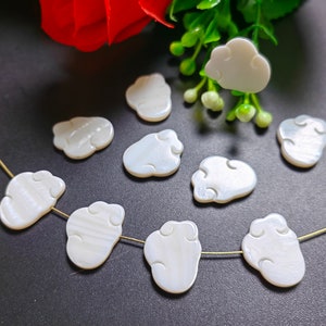 Op de afbeelding: Een draad van witte schelpvormige wolkvormige kralen met een gouden draad erdoorheen geregen. De kralen zijn in een lijn op een zwart oppervlak gerangschikt.