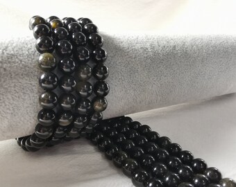 May include: A strand of black gemstone beads with a golden sheen. The beads are round and smooth, and they are strung together on a clear thread. The beads are arranged in a spiral pattern, and they are lying on a white surface.