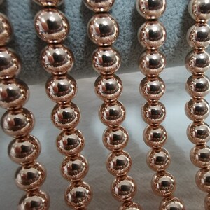 May include: Close-up of several strands of rose gold beads. Each bead is a perfect sphere, reflecting light and creating a shiny, metallic appearance. The beads are strung together, suggesting they are part of a necklace or bracelet.