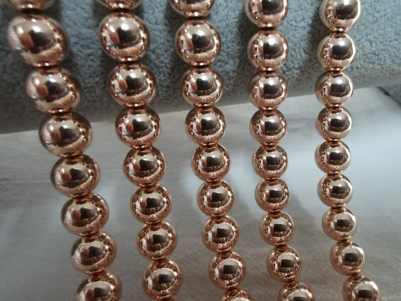 May include: Close-up of several strands of rose gold beads. Each bead is a perfect sphere, reflecting light and creating a shiny, metallic appearance. The beads are strung together, suggesting they are part of a necklace or bracelet.