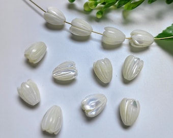 May include: Iridescent white tulip-shaped beads. Some beads are strung on a gold wire, while others are loose. The beads have a pearlescent sheen and are arranged on a white surface with green foliage in the background. These beads are suitable for jewelry making.