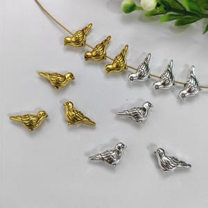 Könnte beinhalten: Eine Sammlung kleiner, vogelformiger Anhänger in Gold- und Silbertönen. Einige Anhänger sind auf goldfarbenem Draht aufgefädelt. Die Vögel sind detailliert mit Flügeln, Schnäbeln und Schwanzfedern gestaltet. Ideal für Schmuckherstellung.