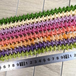 May include: Multiple strands of small, trumpet-shaped flower beads in various colors including green, cream, purple, pink, orange, and yellow, arranged in parallel rows next to a metal ruler showing measurements in centimeters.