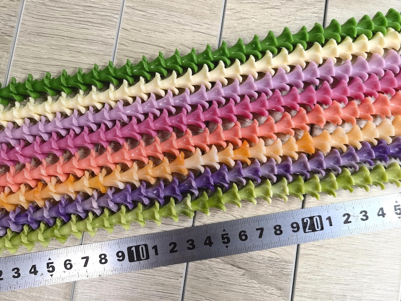May include: Multiple strands of small, trumpet-shaped flower beads in various colors including green, cream, purple, pink, orange, and yellow, arranged in parallel rows next to a metal ruler showing measurements in centimeters.