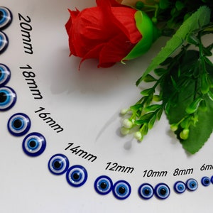 Könnte beinhalten: Sortiment an blauen bösen Augen-Anhängern in verschiedenen Größen von 6 mm bis 20 mm. Die Anhänger sind in einer Reihe auf einer weißen Oberfläche angeordnet, wobei der größte Anhänger oben und der kleinste unten ist.