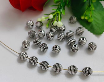 May include: A collection of silver metal beads with a textured surface. The beads are arranged in a circle on a white surface. Some of the beads are strung on a thin wire.