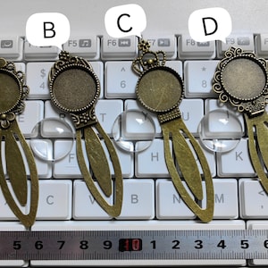 May include: Four bronze bookmark pendants with different designs. Each pendant has a round glass cabochon in the center. The pendants are displayed on a white keyboard with a ruler underneath.