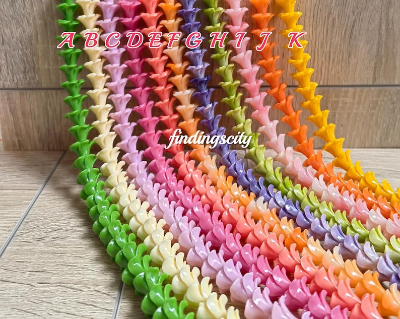 May include: Multiple strands of small, trumpet-shaped flower beads arranged in a vertical gradient of colors including green, cream, pink, orange, purple, and yellow. Each strand is labeled with letters A through K, and the text findingscity is visible.