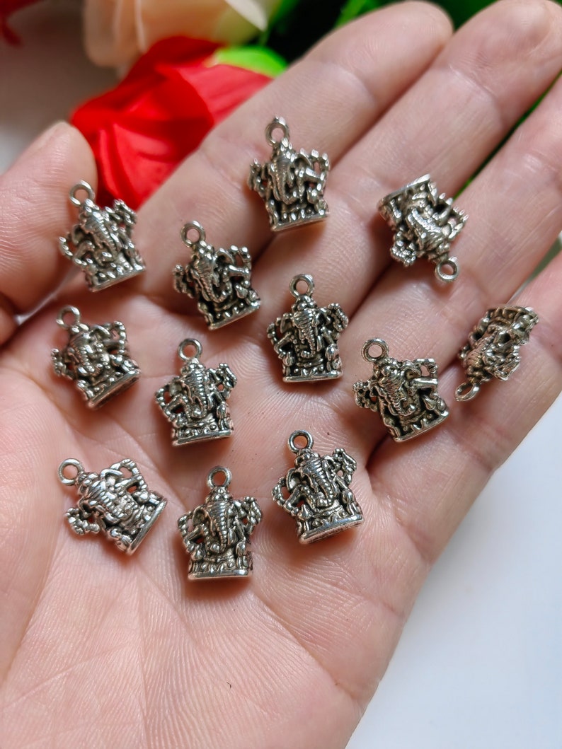 May include: A collection of small, silver-colored charms in the shape of the Hindu deity Ganesha. Each charm features intricate details and a small loop at the top for attaching to jewelry or other crafts. The charms are displayed against a neutral background.