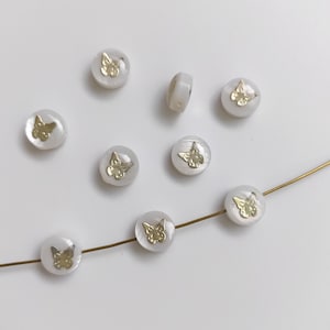 Könnte beinhalten: Eine Schnur aus weißen Perlen mit goldenen Schmetterlingsakzenten. Die Perlen sind auf einem goldenen Draht aufgefädelt.