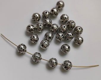 May include: A collection of silver metal beads with intricate designs. The beads are scattered on a white surface and some are strung on a thin wire.