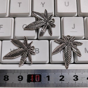 May include: Three silver-toned metal charms shaped like marijuana leaves. The charms are on a white keyboard with black keys and a ruler with black markings.