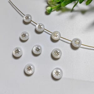 May include: A strand of white beads with gold star accents. The beads are strung on a gold wire and are arranged in a circle.