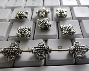 May include: Silver cross-shaped beads with a textured surface. The beads are arranged on a white keyboard.