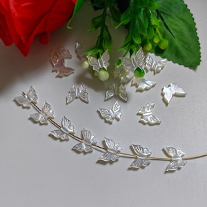 May include: A string of white mother of pearl butterfly beads on a gold wire. The beads are arranged in a curved line, with the wings of the butterflies facing outwards. There are also several loose butterfly beads scattered around the string.