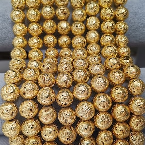 Op de afbeelding: Close-up van talrijke goudkleurige, getextureerde kralen. De bolvormige kralen hebben een ruw, putjesoppervlak. De opstelling suggereert dat ze deel uitmaken van een ketting of armband, wat een sieradenontwerp laat zien.