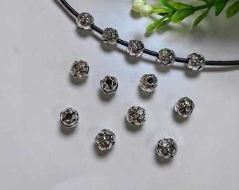 May include: Silver-colored metal beads with a floral design, some strung on a black cord. Several loose beads are scattered on a white surface. Green foliage is visible in the background.