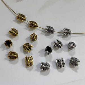 May include: A collection of gold and silver metal bead caps with a leaf design. The bead caps are arranged on a white surface.